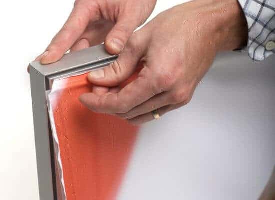 Beursstand Close-up of hands assembling or stretching an orange fabric into a silver frame, possibly for a display or signage. The person is wearing a checked shirt and a gold ring on their left hand.