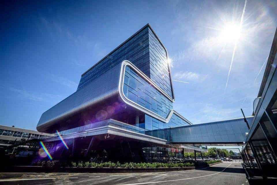 Beursstand A modern glass building with a bold, futuristic design and rounded edges stands under a bright blue sky with the sun shining, lens flare visible, and a skybridge connecting to another structure.