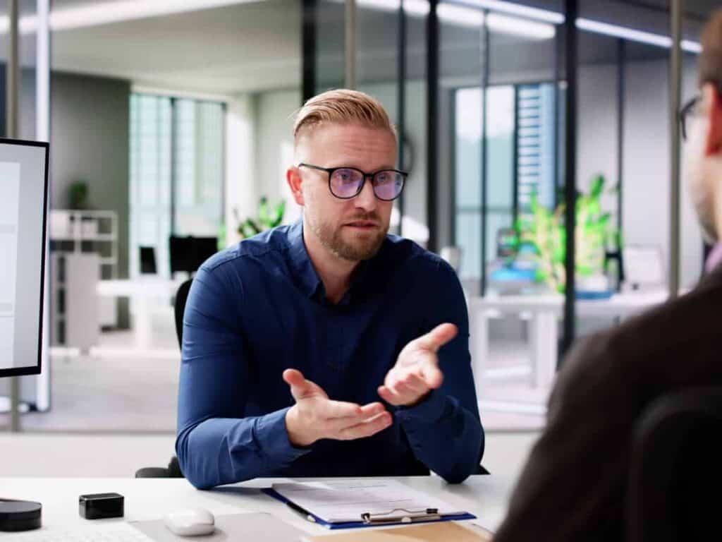 Beursstand Een man met een bril en een blauw shirt zit aan een bureau met documenten en gebaart met zijn handen terwijl hij praat met een andere persoon in een modern kantoor.