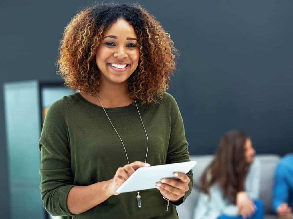 Beursstand Een vrouw met krullend haar en een groene trui lacht terwijl ze een tablet vasthoudt. Ze staat binnen, met twee mensen vaag op de achtergrond, wat een informele of kantooromgeving suggereert.